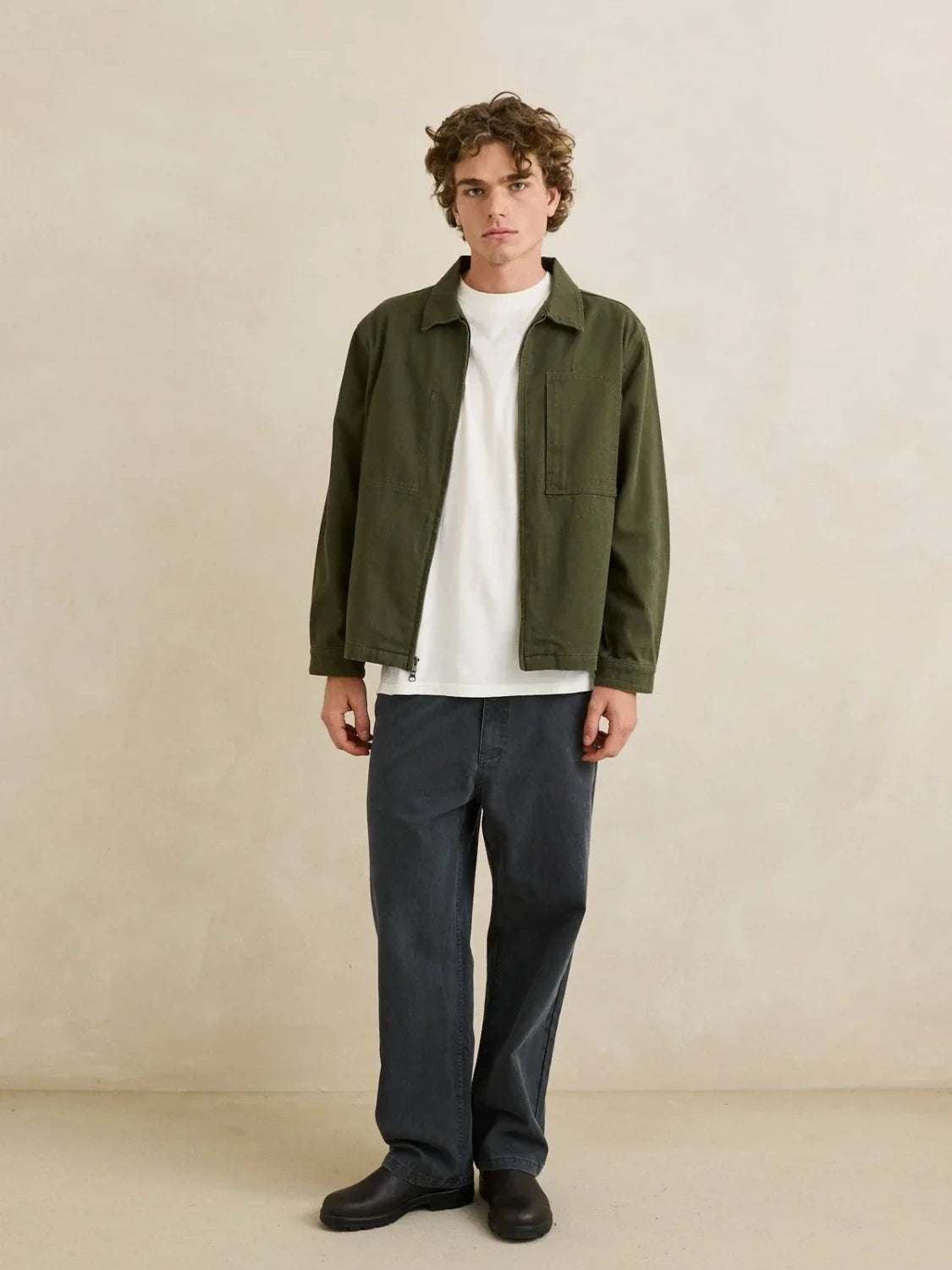 Man wearing a green jacket, white shirt, and dark pants on a beige background