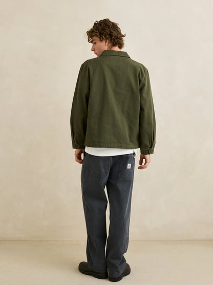 A man is shown from behind wearing an olive green jacket, a white t-shirt, dark blue jeans, and black shoes, standing against a textured beige wall.