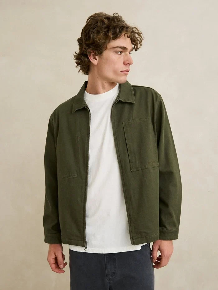 A man wearing an olive green utility jacket over a white t-shirt and dark gray pants, standing against a neutral beige wall.