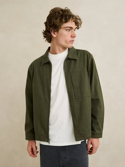A man wearing an olive green utility jacket over a white t-shirt and dark gray pants, standing against a neutral beige wall.