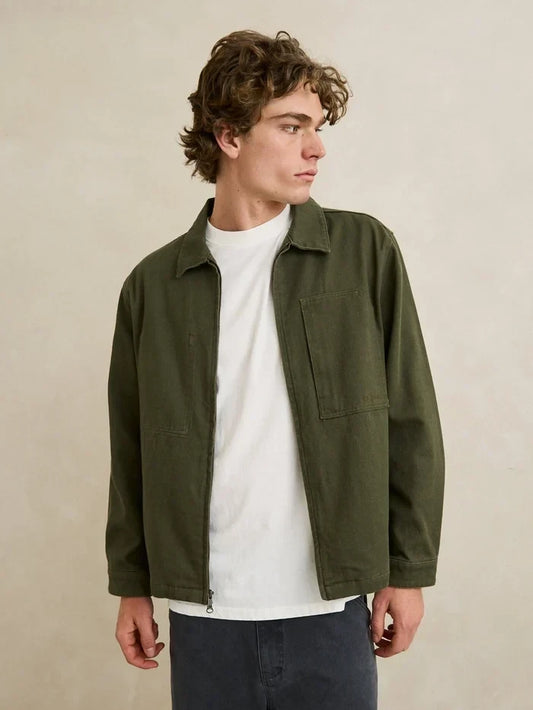 A man wearing an olive green utility jacket over a white t-shirt and dark gray pants, standing against a neutral beige wall.