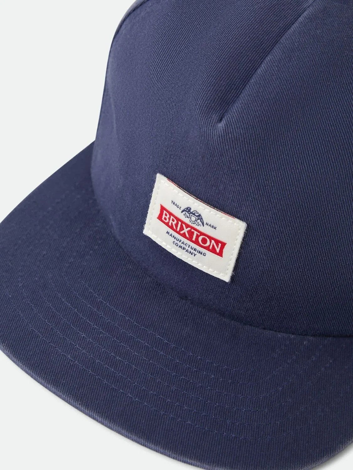 A close-up shot of a navy blue Brixton cap with a white rectangular patch on the front displaying the brand name 'BRIXTON' in red and black text.