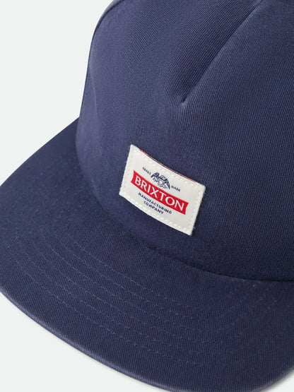 A close-up shot of a navy blue Brixton cap with a white rectangular patch on the front displaying the brand name 'BRIXTON' in red and black text.