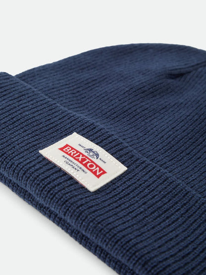 A close-up shot of a navy blue ribbed knit beanie hat, showing a white rectangular Brixton brand patch sewn onto the cuff.