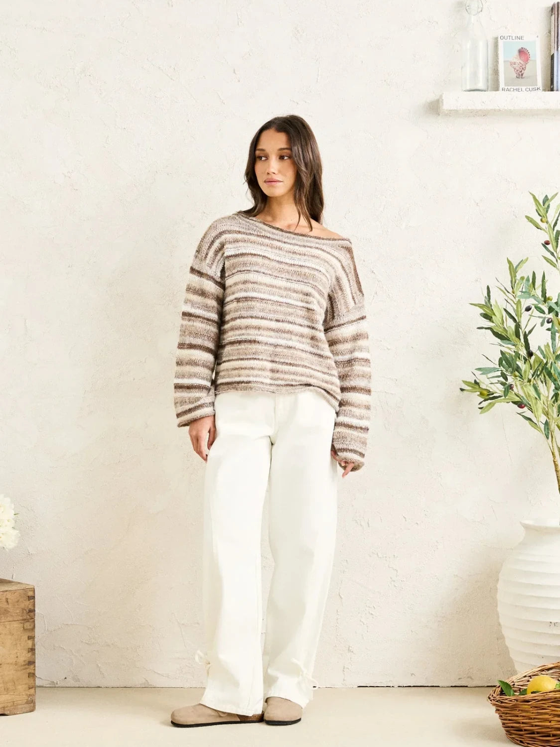 Woman wearing a striped sweater and white pants standing in a room with a plant and basket.