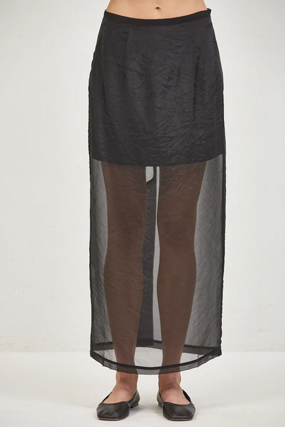 A model wearing a black textured midi skirt with a sheer, floor-length overlay panel, paired with sheer brown tights and black flats, against a plain white studio background.